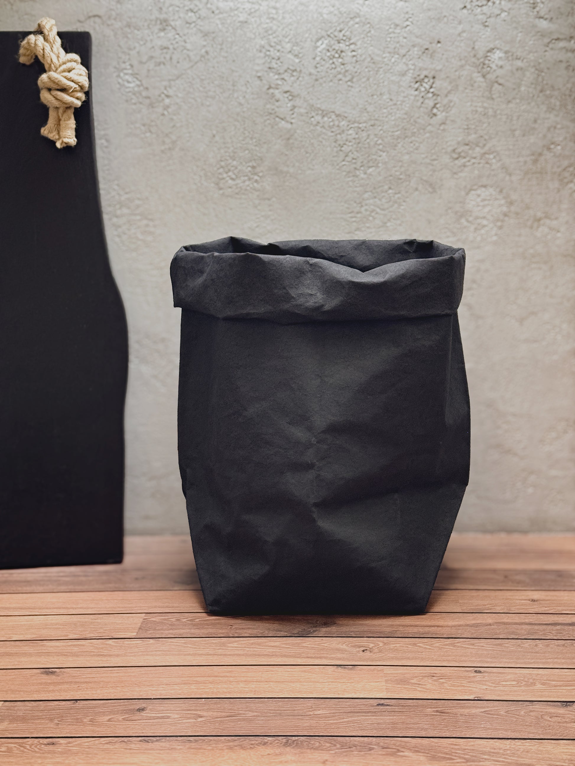 A black washable paper bag with a rolled top, standing on a wooden floor against a textured light gray wall. A black board with a beige rope knot hangs on the left side.