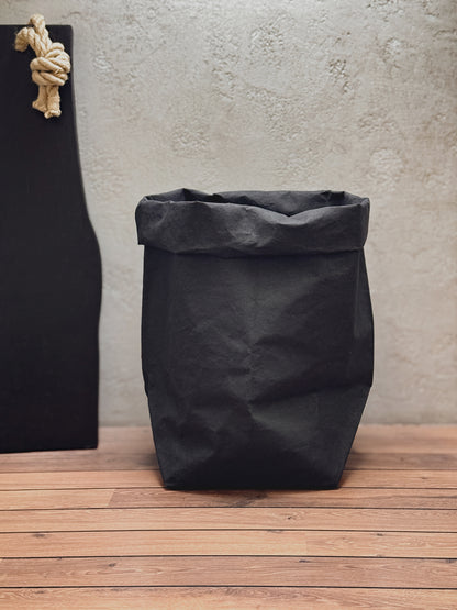 A black washable paper bag with a rolled top, standing on a wooden floor against a textured light gray wall. A black board with a beige rope knot hangs on the left side.