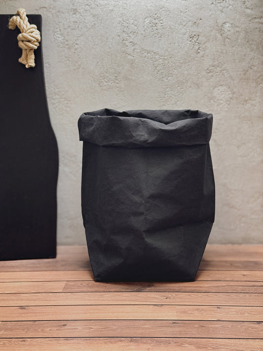 A black washable paper bag with a rolled top, standing on a wooden floor against a textured light gray wall. A black board with a beige rope knot hangs on the left side.