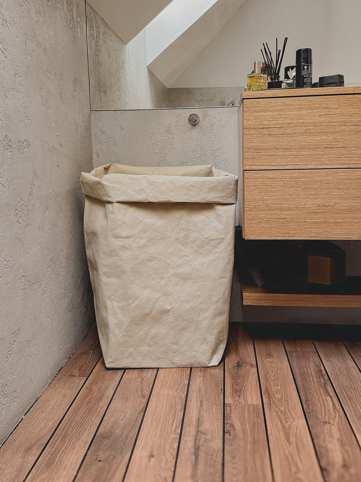 A tall, sand color washable paper laundry hamper bag standing on a wooden plank floor next to a light wood vanity cabinet in a bathroom setting.