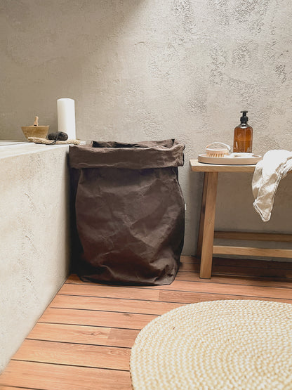 Large washable paper laundry hamper bag standing on a wooden floor next to a bathtub in a rustic bathroom setting.