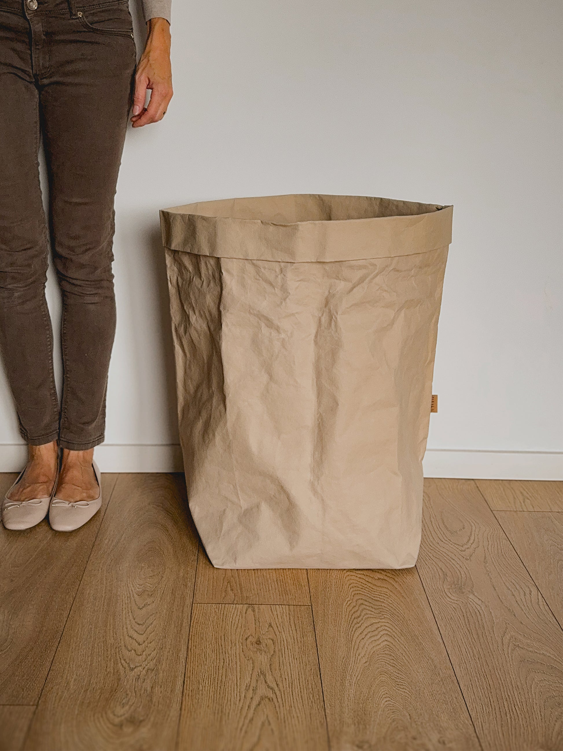 A large, open-top crinkled paper laundry hamper in a natural beige color, standing on a light wood floor against a white wall, with a person's legs visible on the left side to show the bag size