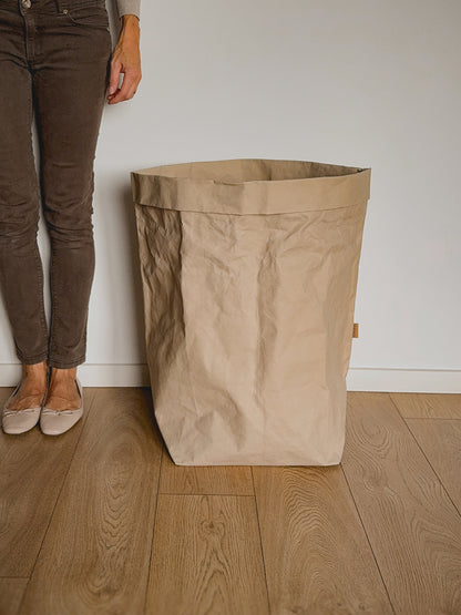 A large, open-top crinkled paper laundry hamper in a natural beige color, standing on a light wood floor against a white wall, with a person's legs visible on the left side to show the bag size