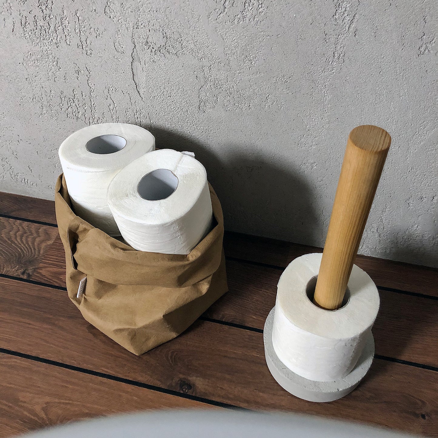 Two rolls of toilet paper in a brown washable paper bag and a concrete holder with a wooden stick on a wooden surface.