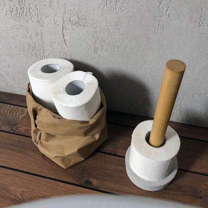 Two rolls of toilet paper in a brown washable paper bag and a concrete holder with a wooden stick on a wooden surface.