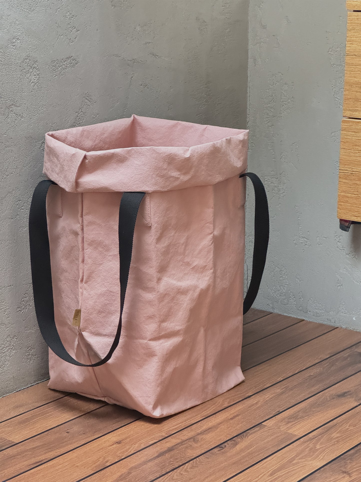 A rose washable paper toy storage bag with black shoulder straps, standing on a wooden deck against a textured gray wall.