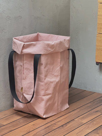 A rose washable paper toy storage bag with black shoulder straps, standing on a wooden deck against a textured gray wall.