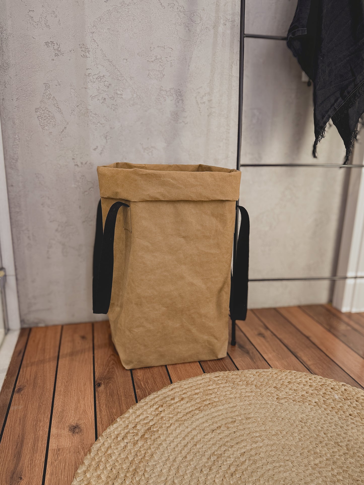 A natural beige washable paper laundry hamper with black fabric handles, standing on a wooden plank floor against a textured light gray concrete wall.