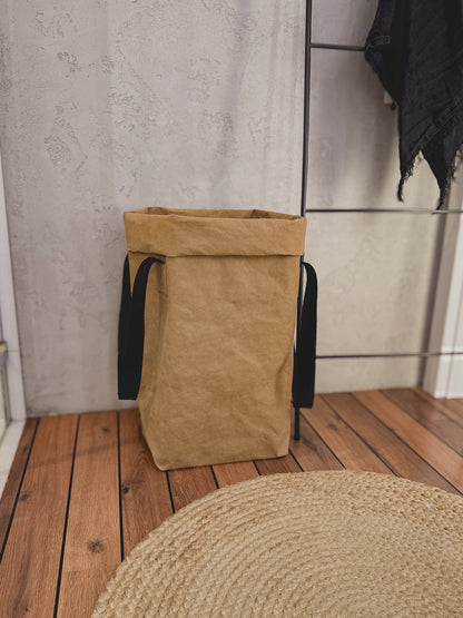 A natural beige washable paper laundry hamper with black fabric handles, standing on a wooden plank floor against a textured light gray concrete wall.