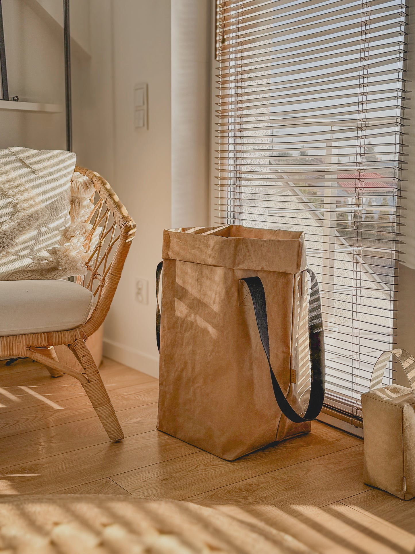 A brown washable paper storage bag with long black handles stands on a wooden floor next to a window with blinds in a sunlit room.