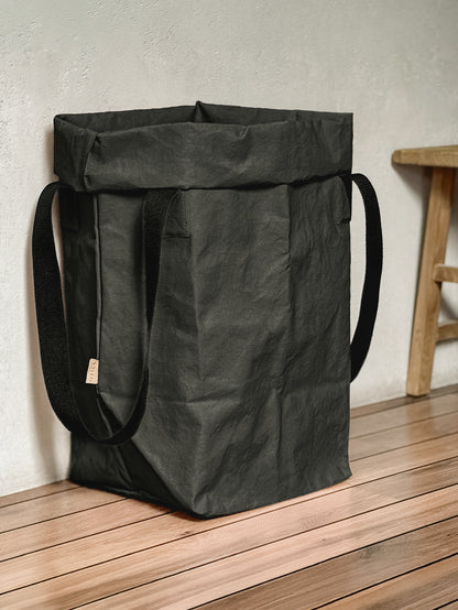 A black washable paper storage bin or laundry hamper with two shoulder straps, standing on a wooden floor against a textured concrete wall.