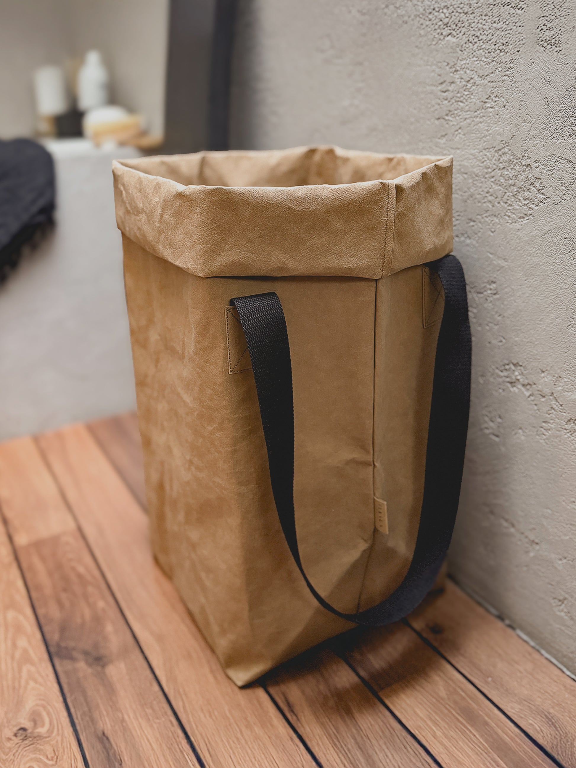A brown washable paper laundry bag with black fabric handles, standing on a wooden deck floor against a textured concrete wall.