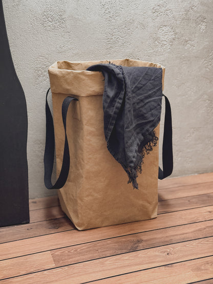 A large brown washable paper laundry hamper with long black shoulder straps, standing on a wooden floor against a textured light concrete wall.
