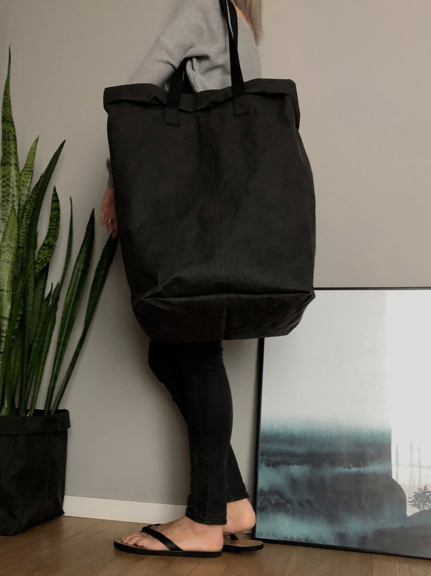 A person carries a large black washable paper laundry basket with long straps on a shoulder in an indoor setting with a potted snake plant in a black paper planter.