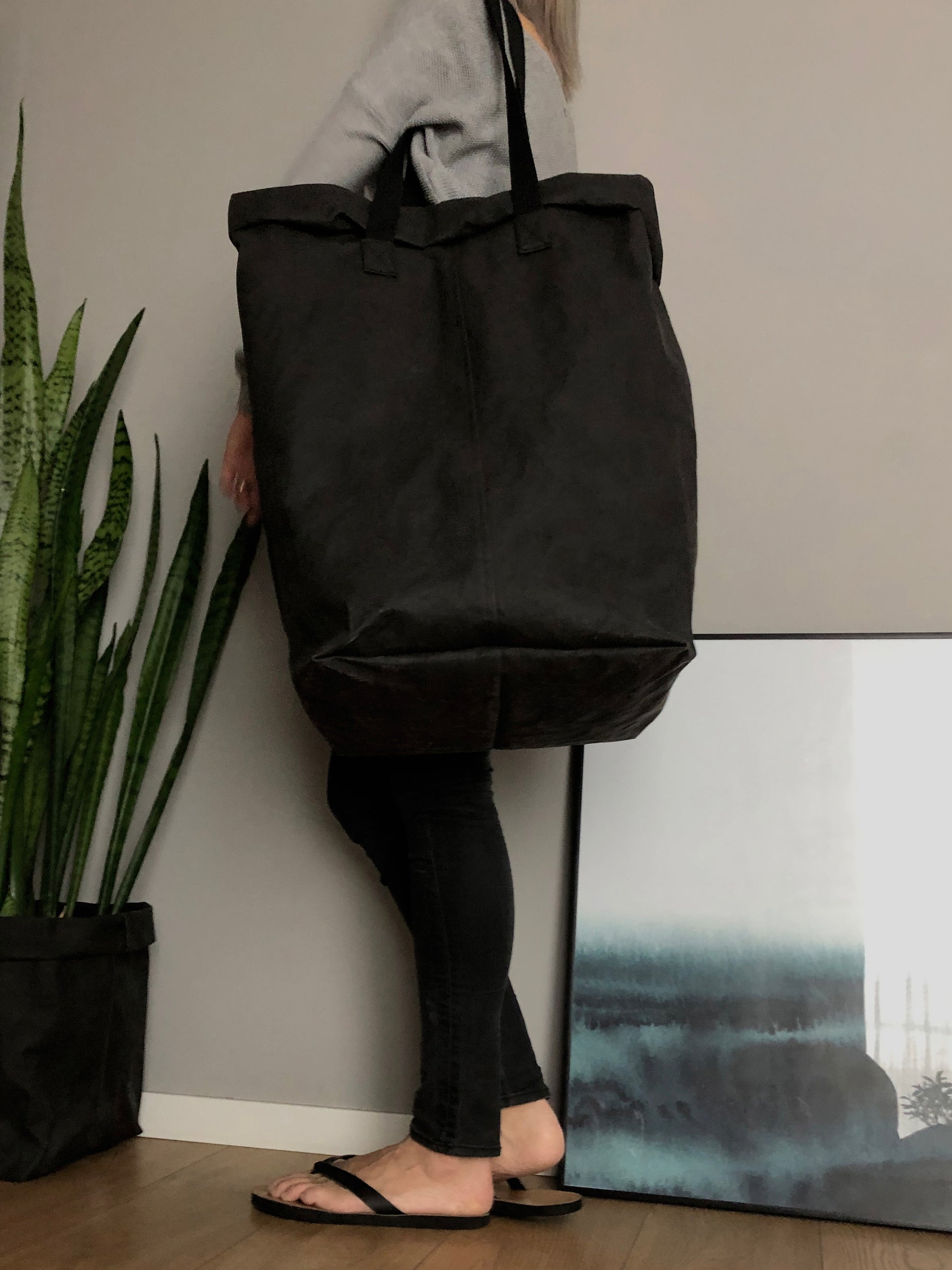 A person carries a large black washable paper laundry basket with long straps on a shoulder in an indoor setting with a potted snake plant in a black paper planter.