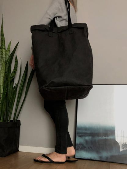 A person carries a large black washable paper laundry basket with long straps on a shoulder in an indoor setting with a potted snake plant in a black paper planter.