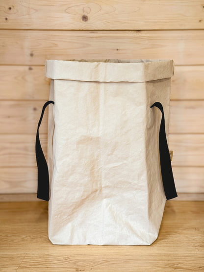 White washable paper laundry basket with black cotton handles against a light wood wall