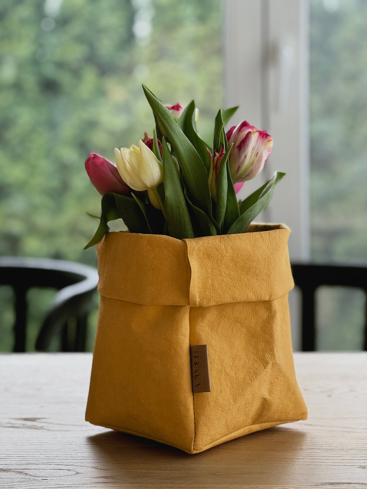A mustard yellow washable paper planter filled with tulips and displayed on a wooden table in front of a softly blurred window.