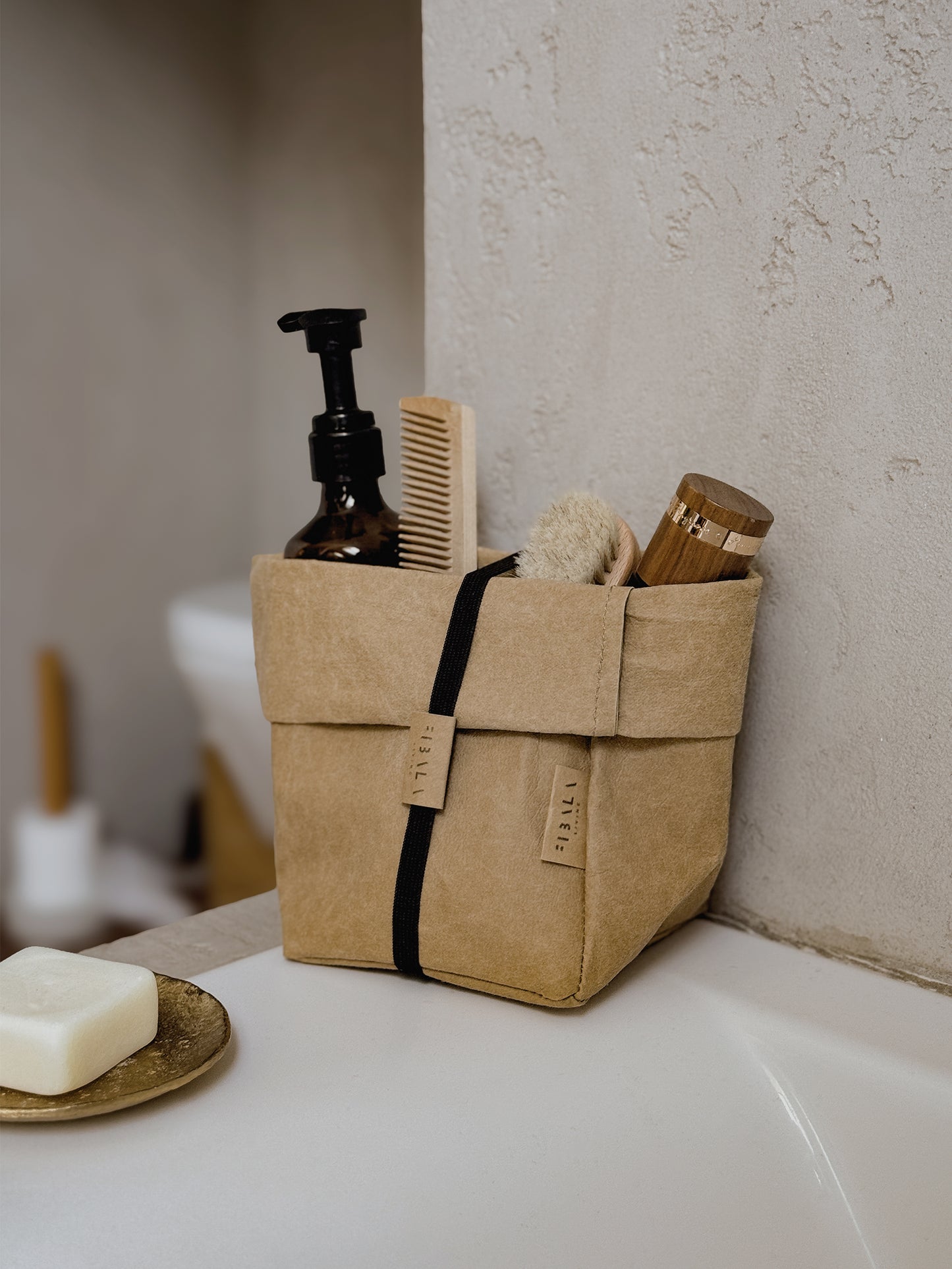 A brown washable paper bathroom organizer basket with a black elastic band, holding cosmetics, a comb and brushes on a bath edge.