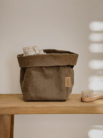 A brown washable paper storage basket with a folded top edge, shown on a wooden table with a small wooden brush beside it against a warm neutral wall.