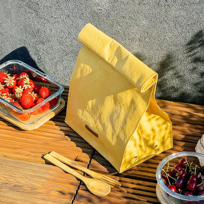 Top-side view of yellow washable paper lunch bag with folded top and metal snap closure, styled with fruit containers on wooden bench and concrete wall backdrop