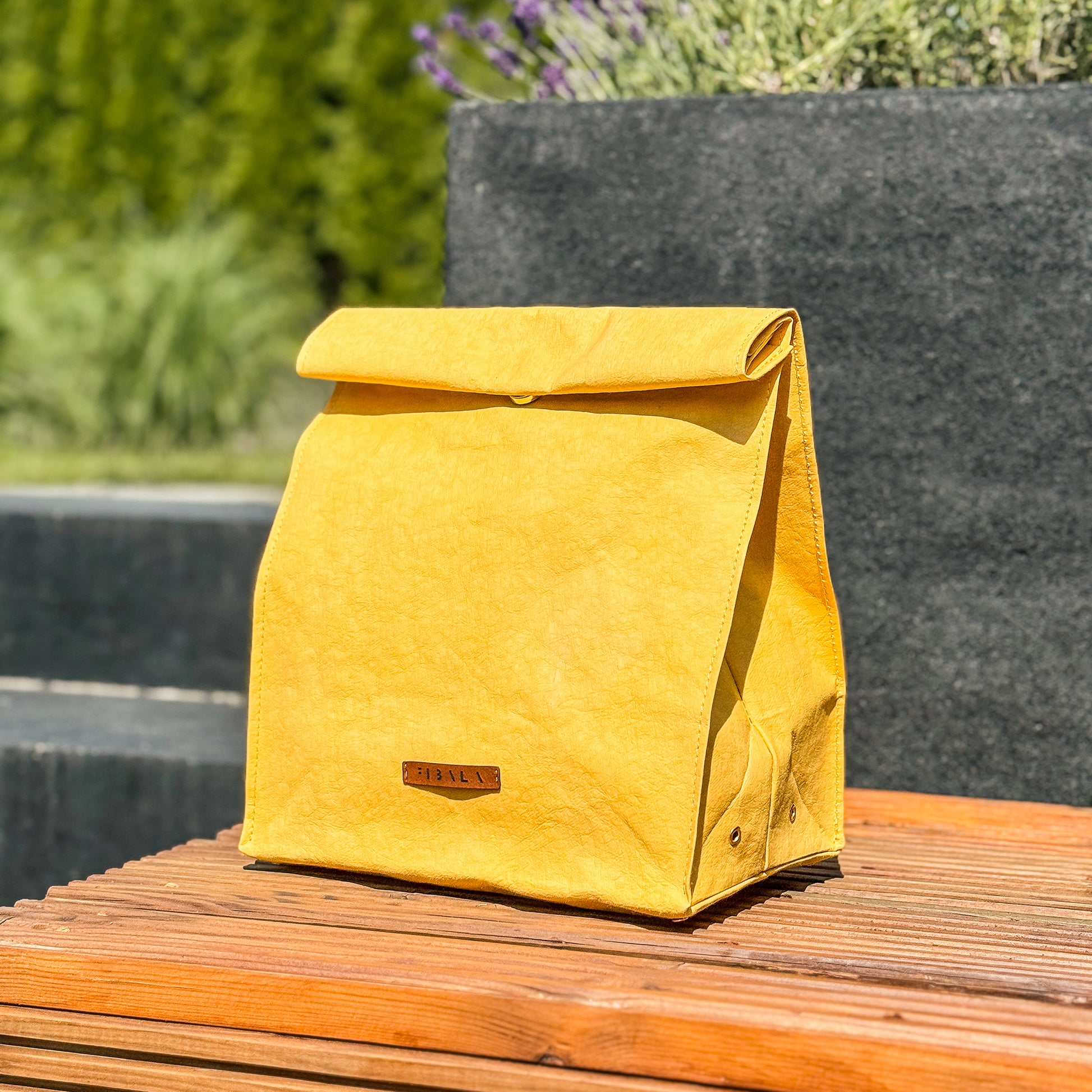 Front view of yellow washable paper lunch bag with folded top and snap closure, standing on wooden bench with concrete planters and garden background.