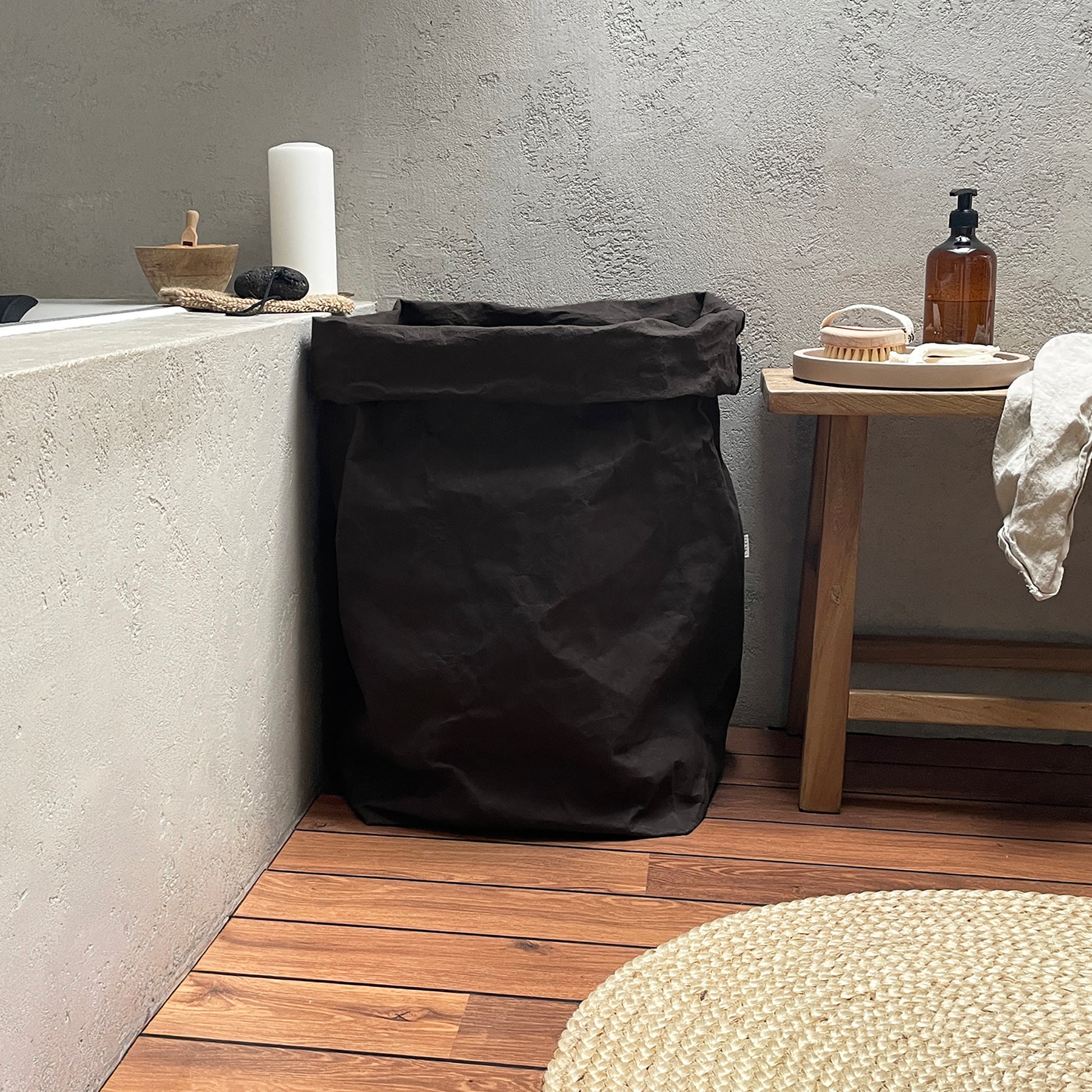 Black washable paper laundry bag in a bathroom setting with a wooden shelf and concrete wall.