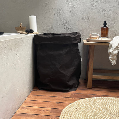 Black washable paper laundry bag in a bathroom setting with a wooden shelf and concrete wall.