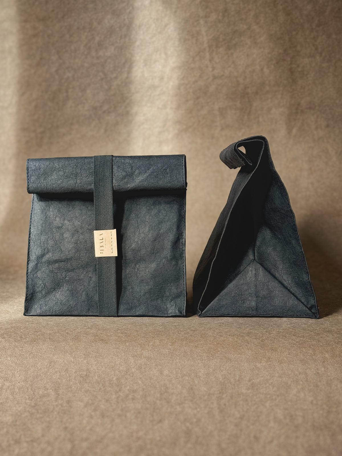 Black washable paper lunch bag with a rolled top and elastic band, shown from the front and the side