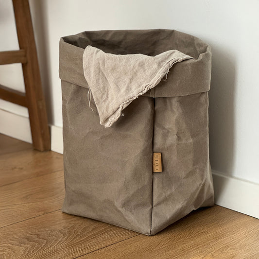 An extra-large washable paper bag in chino brown, featuring a label, placed on a wooden floor against a white wall | Reusable paper bag