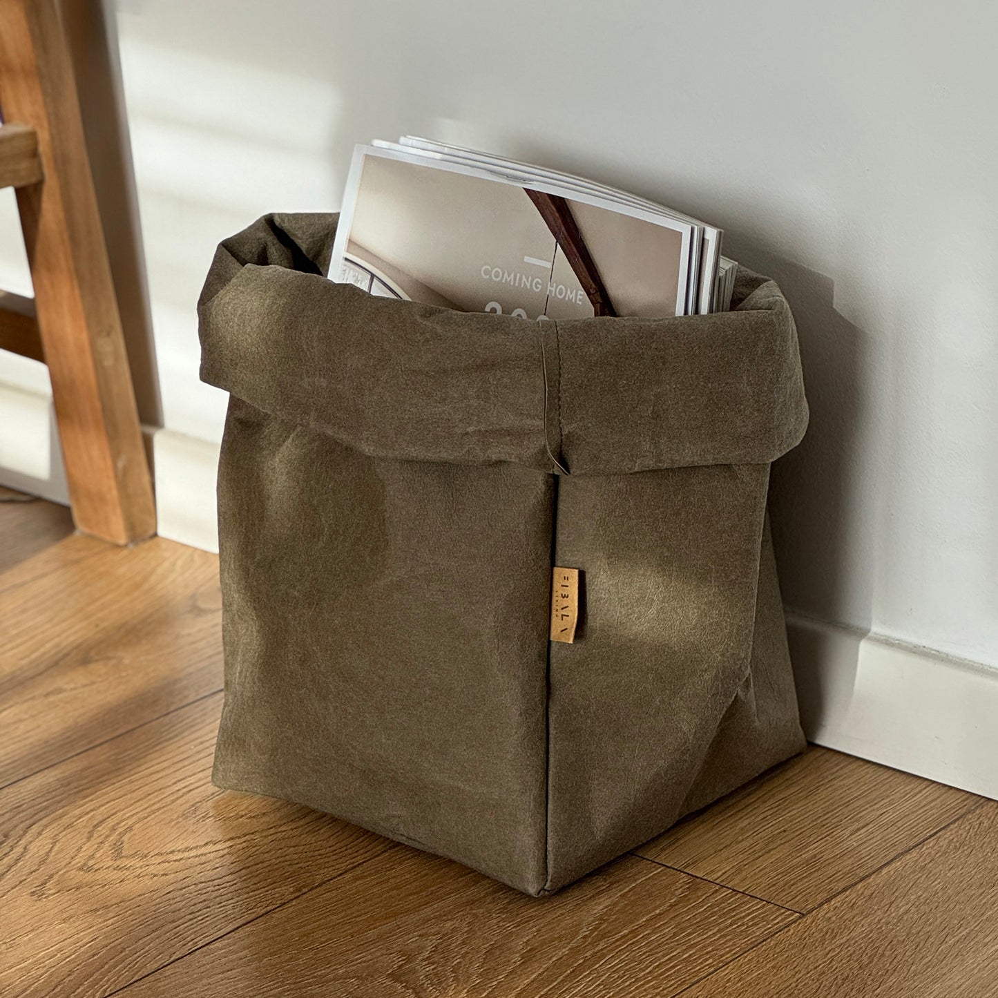Extra-large olive green washable paper bag filled with magazines, on wooden floor, white wall background | Eco-friendly, versatile reusable storage bag