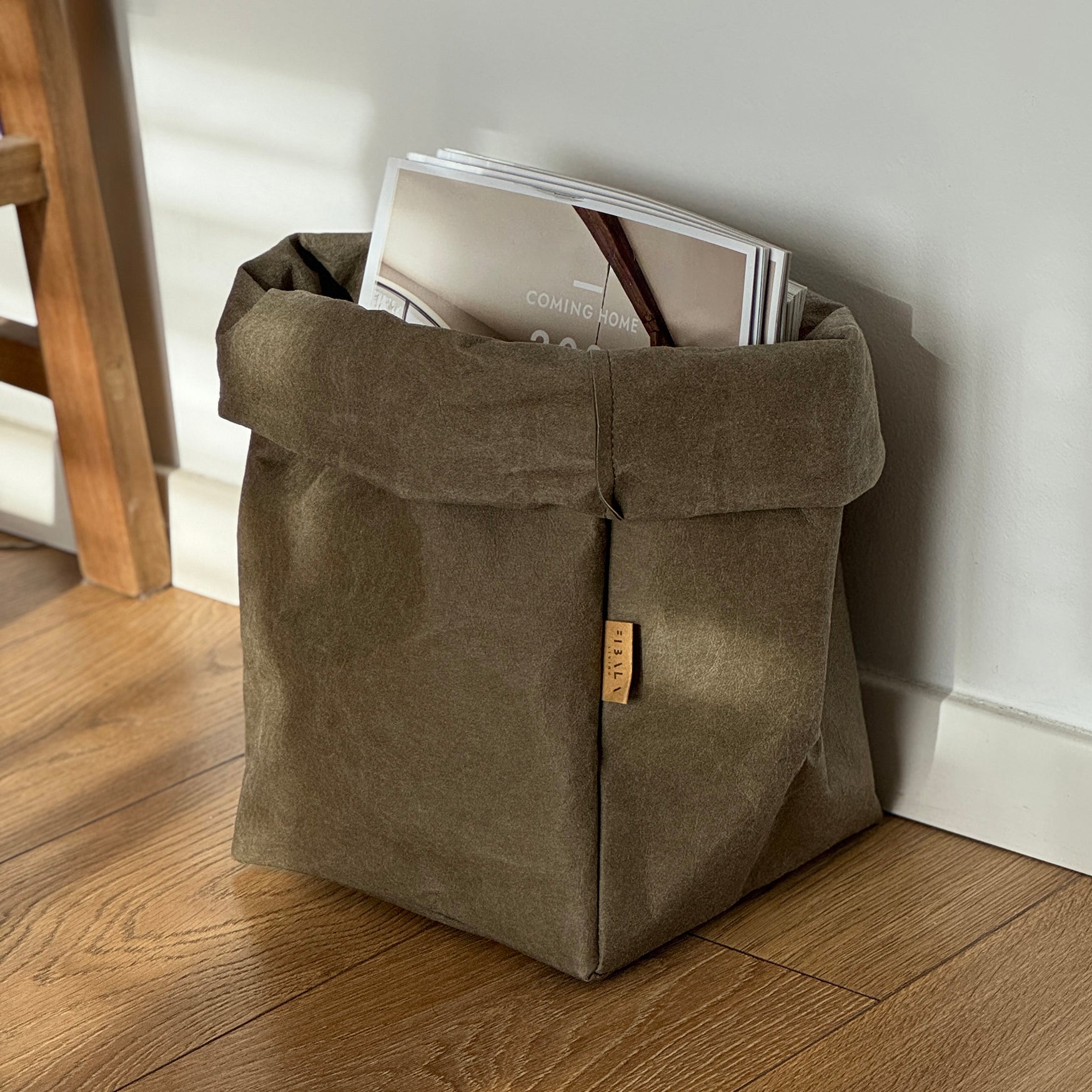 Extra-large olive green washable paper bag filled with magazines, on wooden floor, white wall background | Eco-friendly, versatile reusable storage bag
