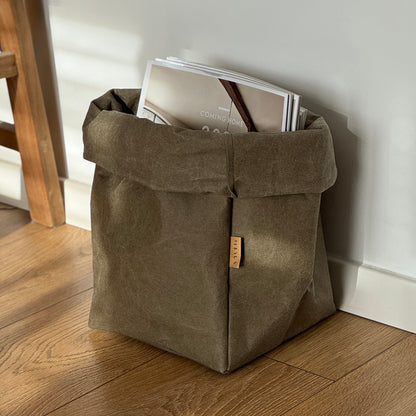 Extra-large olive green washable paper bag filled with magazines, on wooden floor, white wall background | Eco-friendly, versatile reusable storage bag