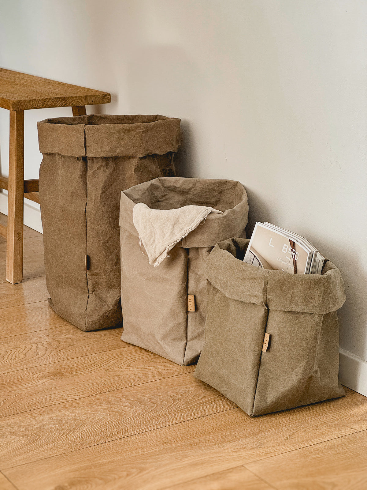 Three washable paper storage bags in different sizes, one holding magazines, by a wooden bench