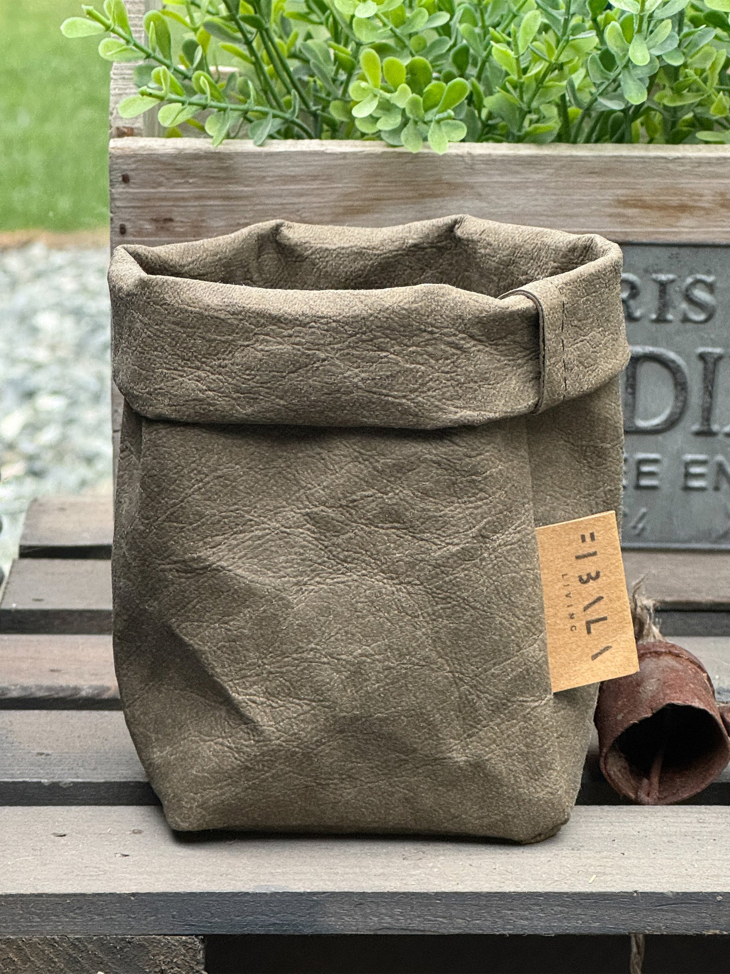 Olive washable paper bag with a visible brand tag on a wooden surface with plants in the background