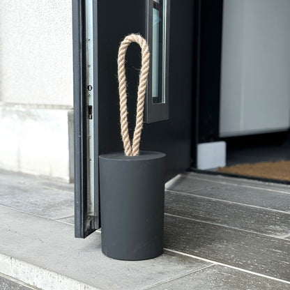Extra large black concrete door stop with a loop-style natural jute rope handle, positioned beside an open exterior door. The close-up highlights the textured concrete surface and twisted rope detail, designed for easy lifting. A bold, functional accent for modern, minimalist spaces and stylish exterior.