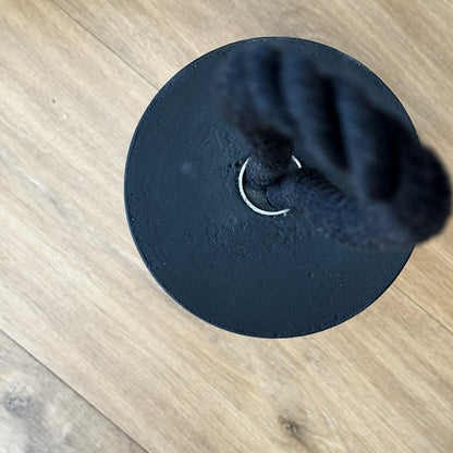 Top-down view of an extra large black concrete door stop, showcasing its smooth cylindrical shape and solid concrete base. The black cotton rope handle appears slightly blurred in the background, keeping focus on the raw texture and clean lines of the concrete. A sleek, functional accent for modern minimalist interiors and exteriors.