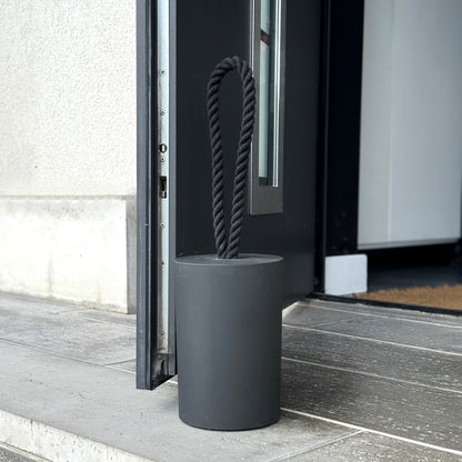 Front view of an extra large black concrete door stop with a loop-style black cotton rope handle, placed next to a modern exterior door and holding it open. The scene features black and grey tones, highlighting the minimalist design and sturdy presence of the door stop. A practical and stylish solution for modern exterior spaces.