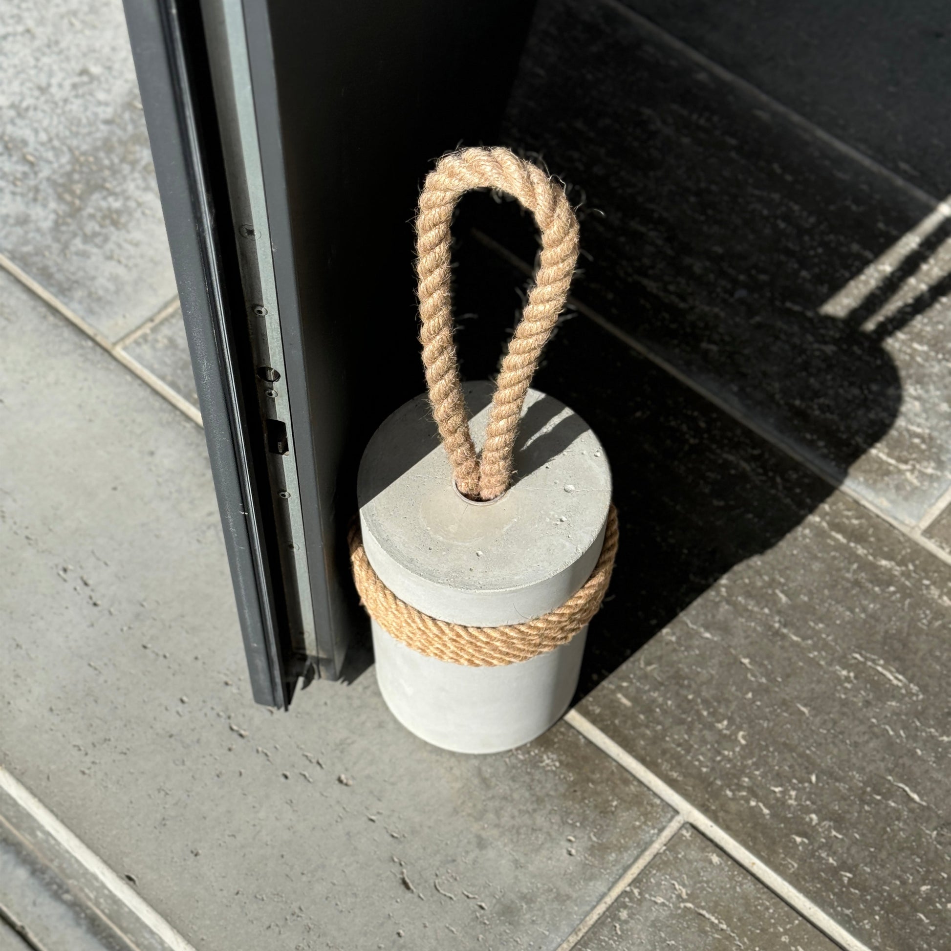 top view of minimalist gray concrete door stop with jute handle and rustic wrap, placed near a patio door on stone flooring — heavy modern doorstop for indoor and outdoor use