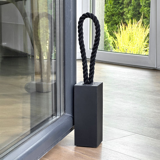 Black concrete door stop with black cotton rope handle, modern rectangular design placed next to patio door inside minimalist living room, garden view in background.