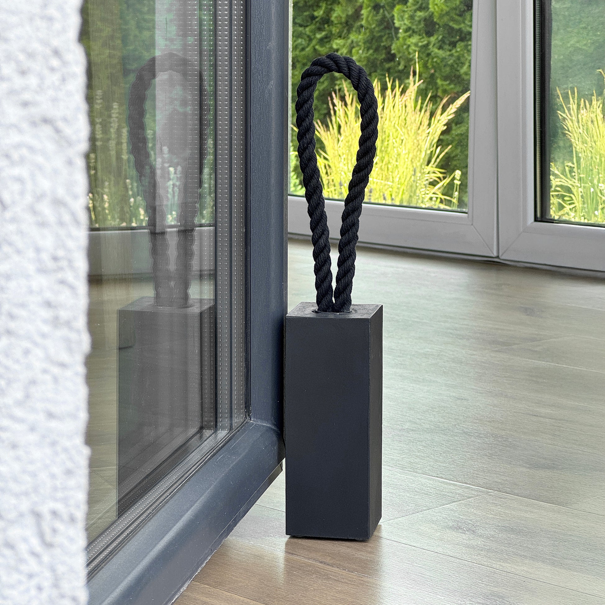 Close-up view of a Black concrete door stop with black cotton rope handle, modern rectangular design placed next to patio door inside minimalist living room, garden view in background.