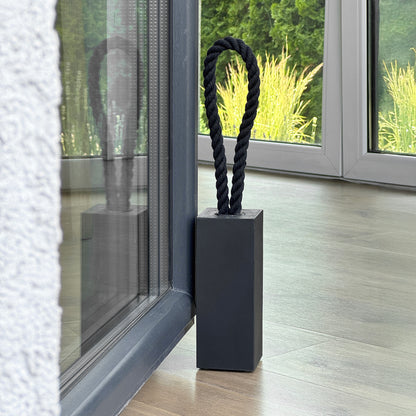 Close-up view of a Black concrete door stop with black cotton rope handle, modern rectangular design placed next to patio door inside minimalist living room, garden view in background.