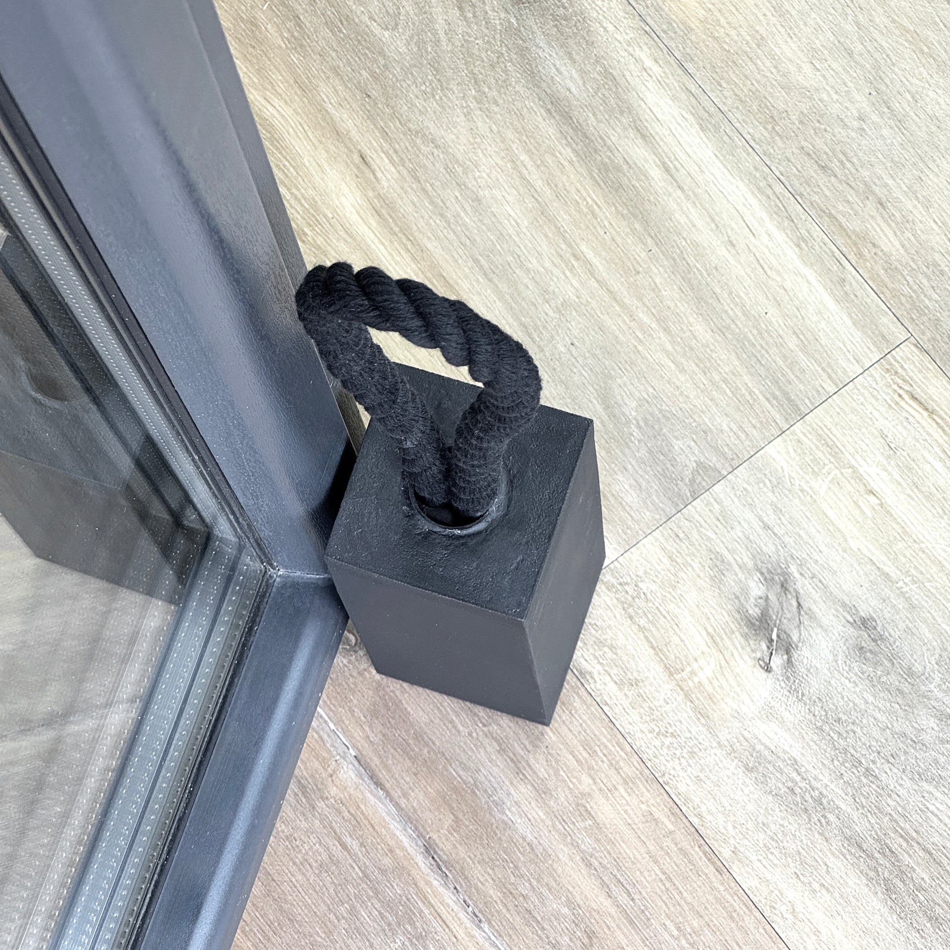 Black concrete door stop in rectangular shape with a black cotton rope handle placed next to a glass door on a wooden floor. Close-up top view.