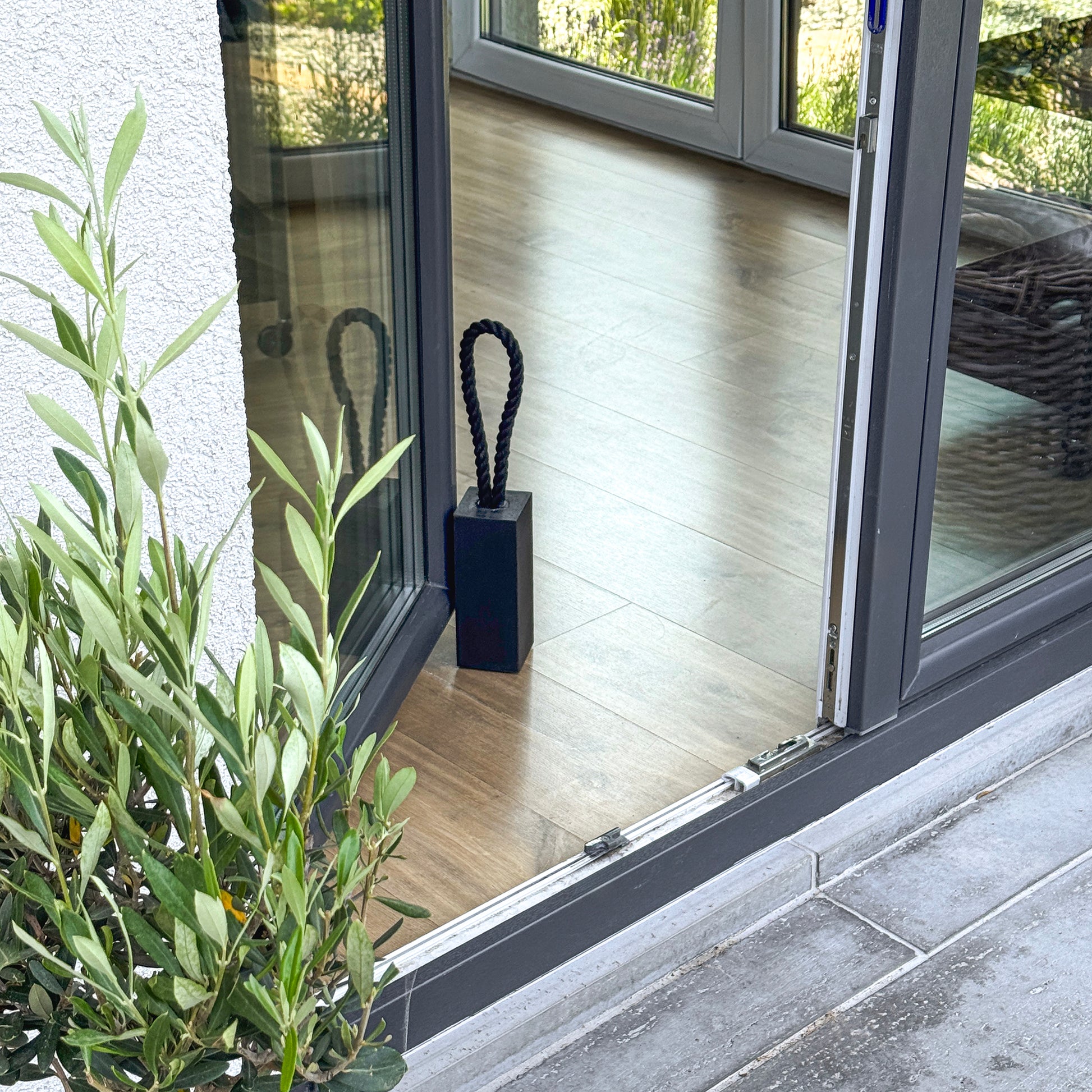 Modern patio door held by a black concrete door stop in rectangular shape with an olive plant in the foreground. 