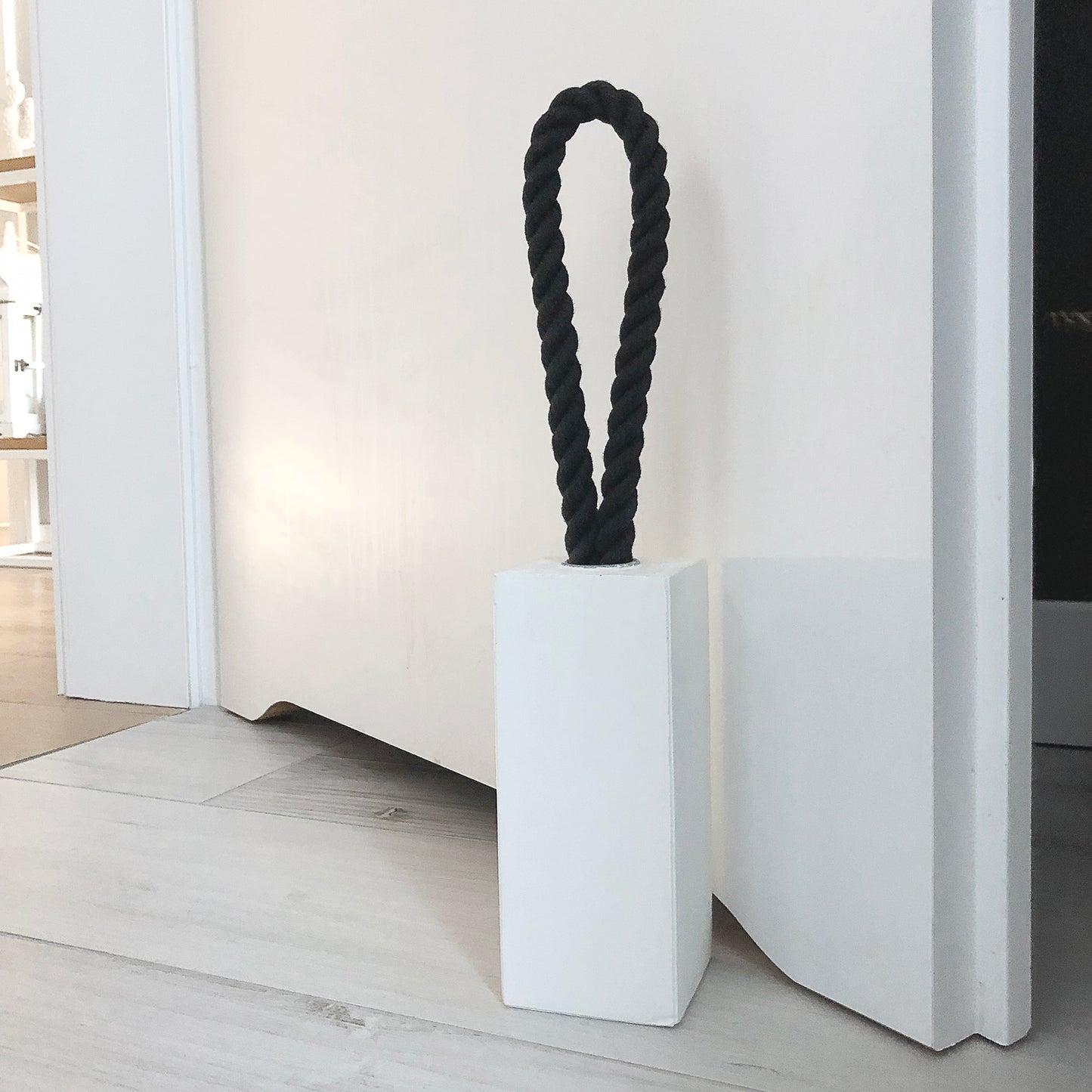 White concrete door stop with a black cotton rope handle, next to white wide open white bathroom door on white tiled floor.