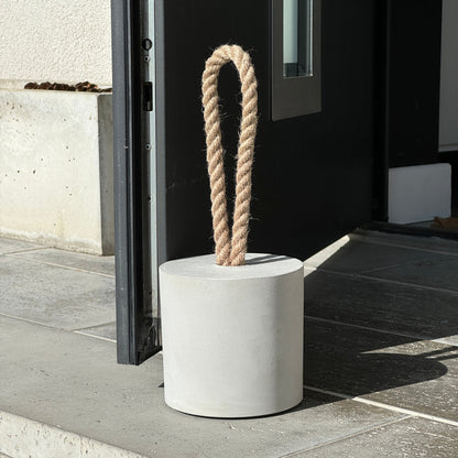 Close-up of an extra-large grey concrete door stopper in a cylindrical shape with a jute handle, holding an exterior door open in a modern outdoor setting. A minimalist, modern door stop designed for outdoor use.
