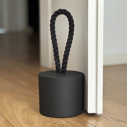 Front view of an extra-large black concrete door stopper with a cylindrical shape and black cotton handle, placed against a door in a neutral modern interior. Sleek and minimalist, ideal for contemporary spaces and suitable for outdoor use.