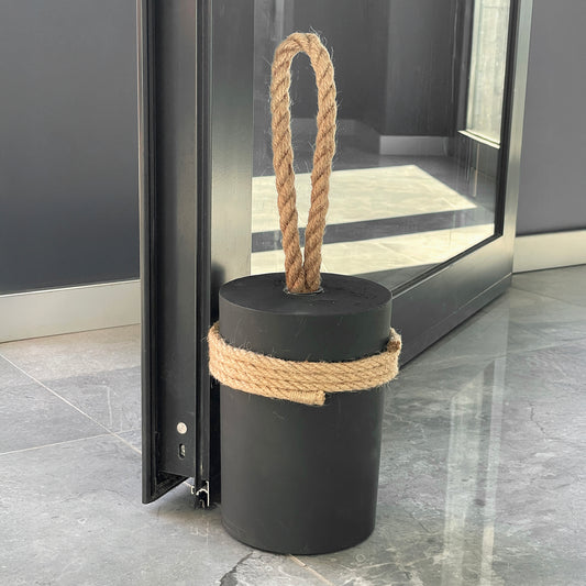 extra-large black concrete door stop with natural jute rope handle and jute string wrap, placed beside a heavy exterior door on marble floor — modern minimalist doorstop for office or hotel entry